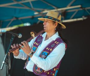 A man in a large hat and traditional style vest plays a flute. 