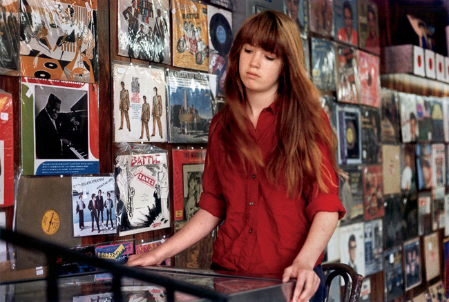 Fotografia colorida de uma garota de camisa vermelha, examinando os registros em uma loja de música.