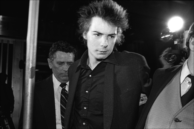 Fotografía en blanco y negro de Sid Vicious. Un hombre a su izquierda lo agarra del brazo y se lo lleva.