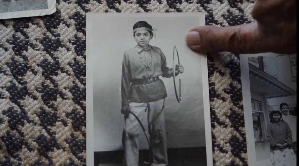 Fotografía en blanco y negro de un joven nativo con un aro en la mano izquierda y otro aro alrededor de la pierna derecha. Un dedo apunta a la fotografía.
