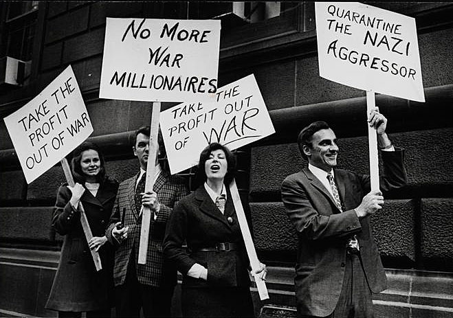 Black and white photograph depicts a group of men and women protesting American involvement in conflicts abroad. Protestors are holding signs addressing their concerns. 