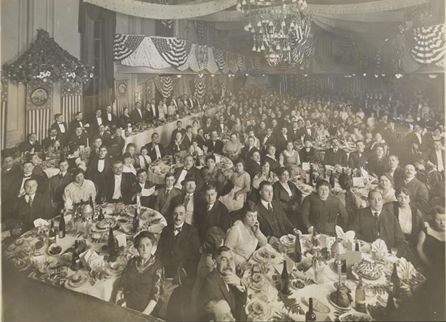 Black and white circa 1910 photograph of a formal dinner banquet. Men, women, and a few children sit at tables looking at camera, place settings, dessert, and wine bottles are visible on the tables.