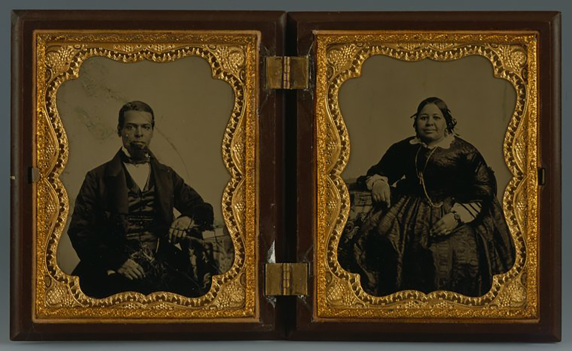 Two black and white photographs in a hinged wood and gold case. The man, left, and woman, right, are black and wear a fashionable dark suit and dress. 