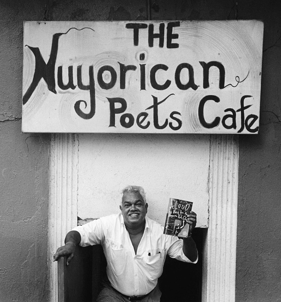 Un hombre, el poeta Miguel Algarín, sonríe y sostiene un libro bajo un cartel que dice: "El Café de los Poetas Nuyorican".