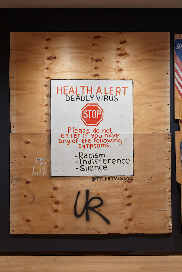 Arte de madeira compensada criada durante a pandemia de COVID-19 e levantes de justiça racial em 2020. Uma placa branca é pintada no centro da madeira com letras vermelhas e pretas e um símbolo de sinal de stop.