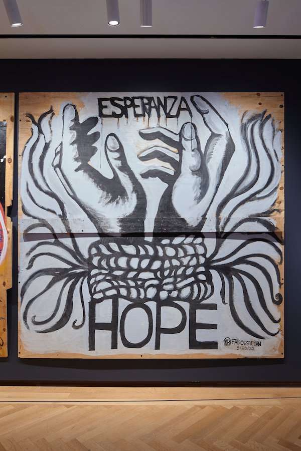 Plywood artwork created during the COVID-19 pandemic and racial justice uprisings in 2020. Bound hands, lined in black against a white background, reach upward. The words "Esperanza" and "Hope" appear above and below. 