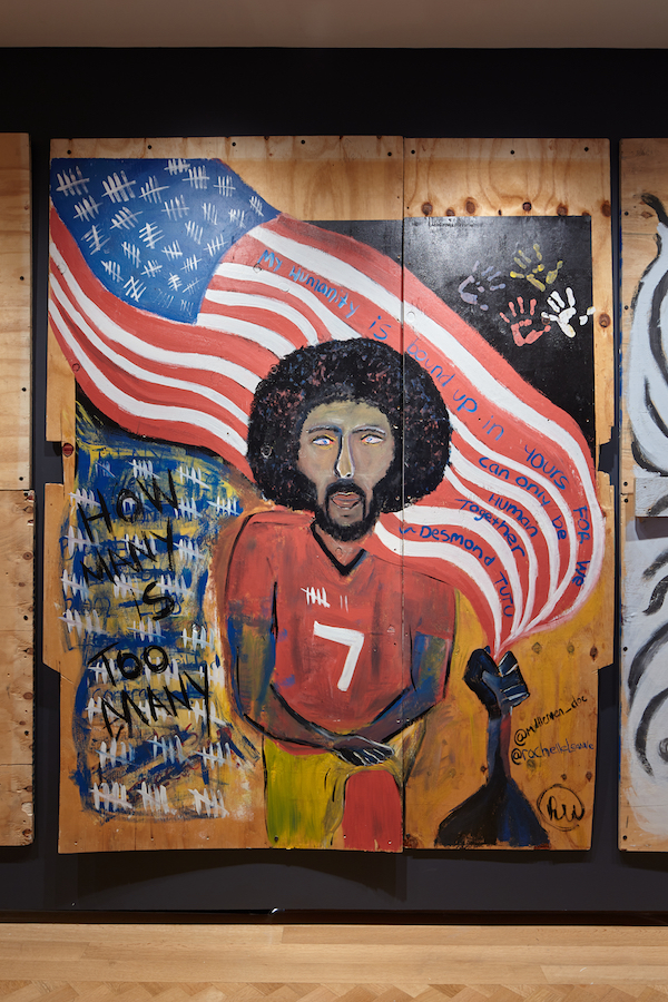 Obras de arte de madera contrachapada creadas durante la pandemia de COVID-19 y los levantamientos por la justicia racial en 2020. Colin Kaepernick está en primer plano, de rodillas, con la bandera estadounidense ondeando detrás de él.