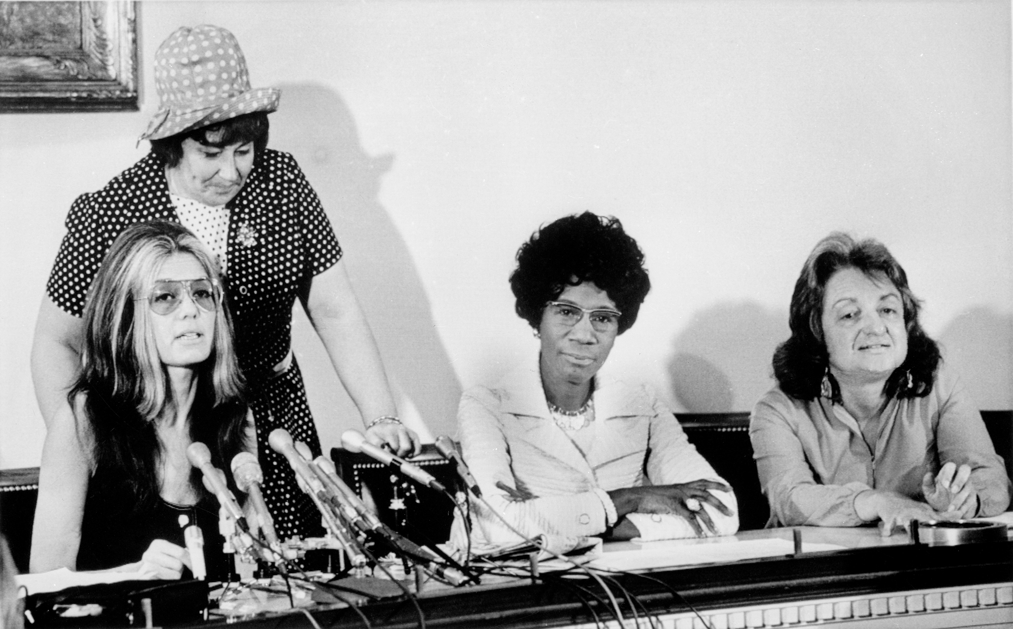 Três mulheres se sentam à mesa, uma com microfones à sua frente, sugerindo que é uma conferência de imprensa