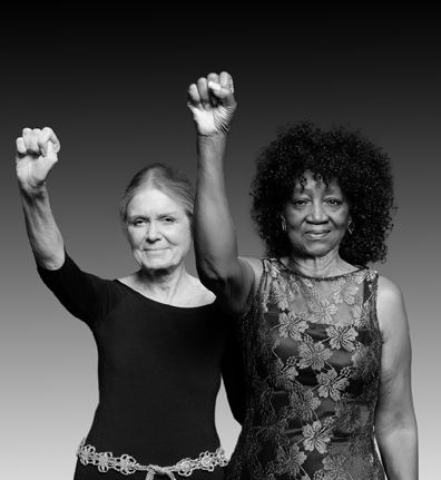 Gloria Steinem e Dorothy Pitman Hughes estão um ao lado do outro, cada um levantando um braço com o punho fechado.