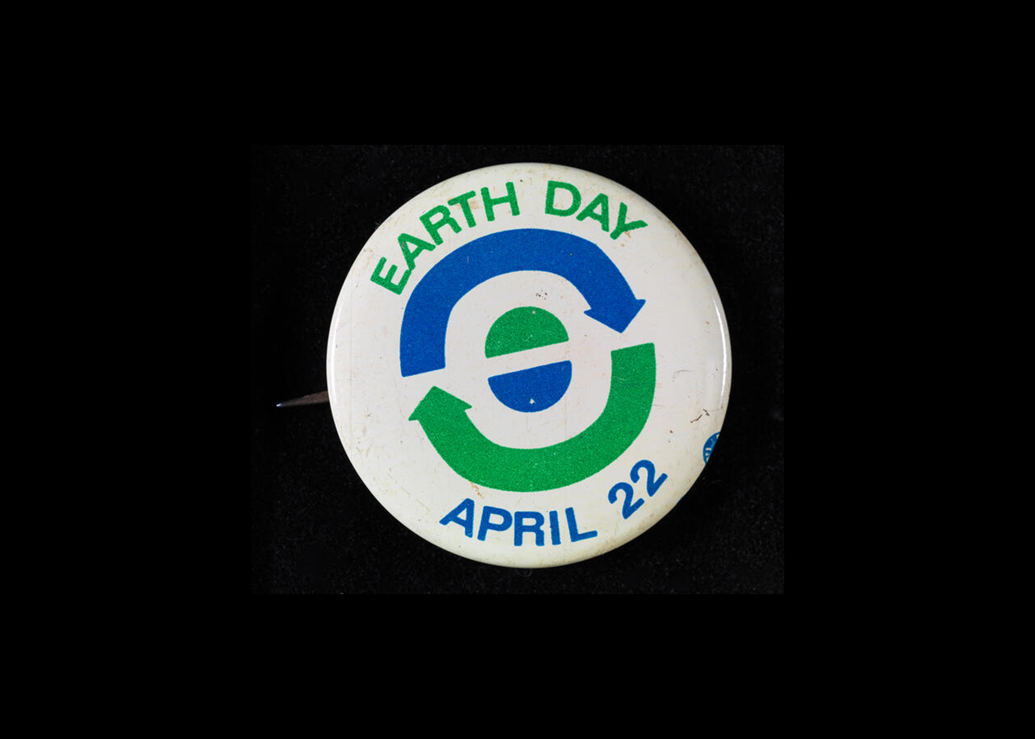 Um botão branco onde se lê Dia da Terra, 22 de abril, com setas azuis e verdes e semicírculos que simbolizam a reciclagem e a terra