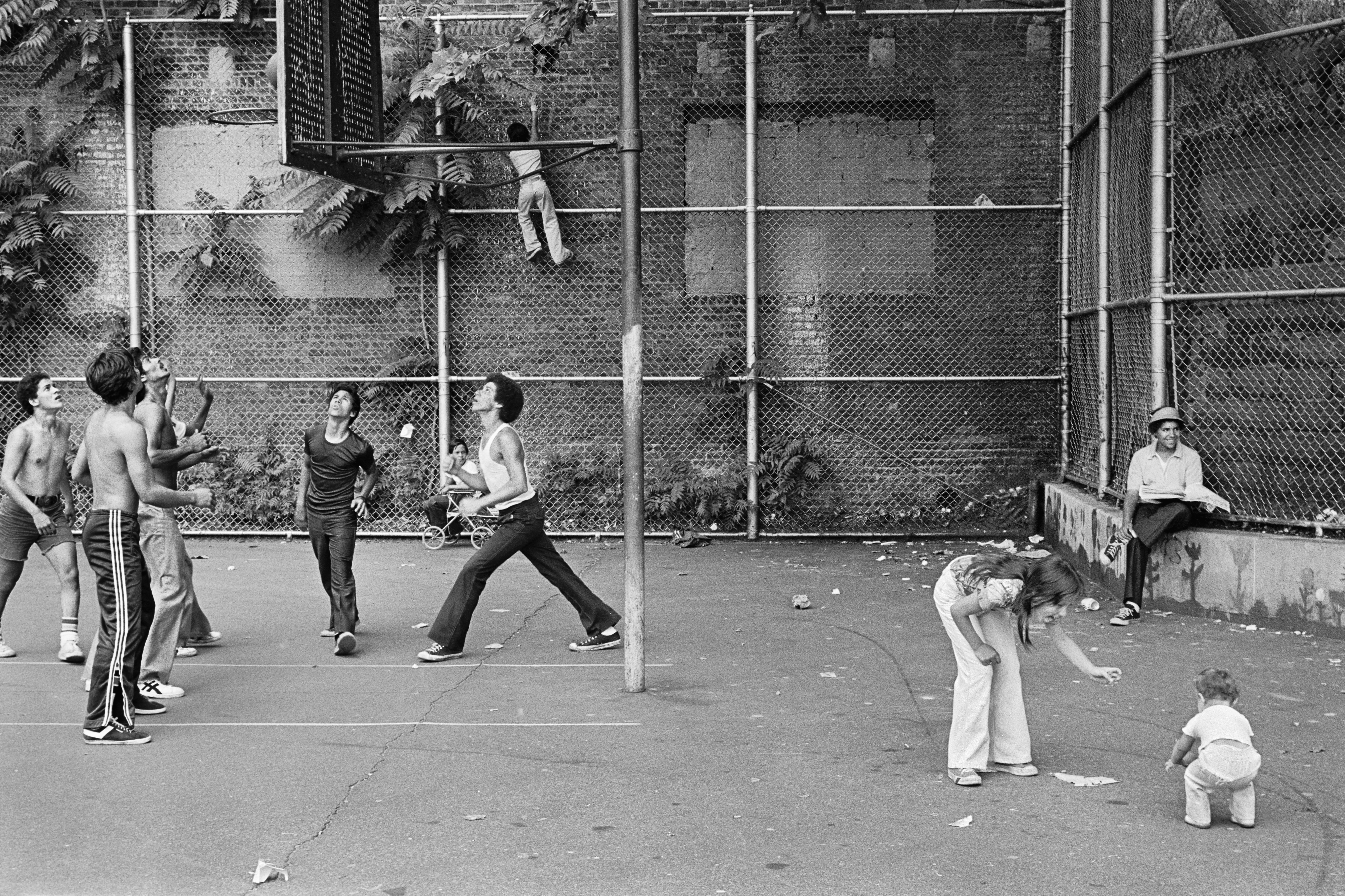 Photographie de personnes jouant au basket-ball dans un terrain de basket entre deux bâtiments, femme et enfant devant