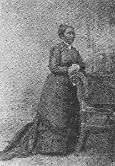 Un retrato de estudio en blanco y negro de cuerpo entero de una mujer negra. Lleva un vestido largo con sobrefalda, bullicio y encaje, y un broche de cuello y aretes.