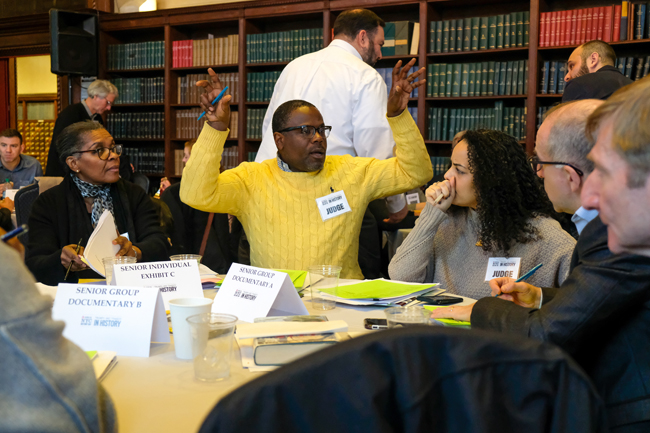 Um grupo de juízes do Dia da História se senta à mesa. Um homem com um suéter amarelo está com os braços levantados, no meio da discussão.