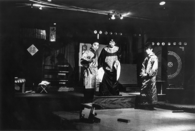 Fotograma de teatro en blanco y negro de "El cuento del abanico románico". Tres actores vestidos con trajes tradicionales chinos se destacan, uno a la izquierda girando hacia la residencia.