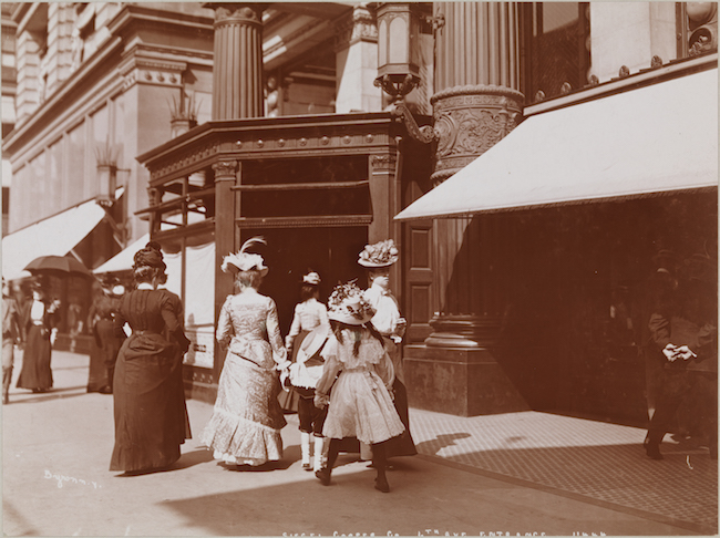Photographie d'un groupe de femmes en robe du XIXe siècle marchant devant un grand magasin.