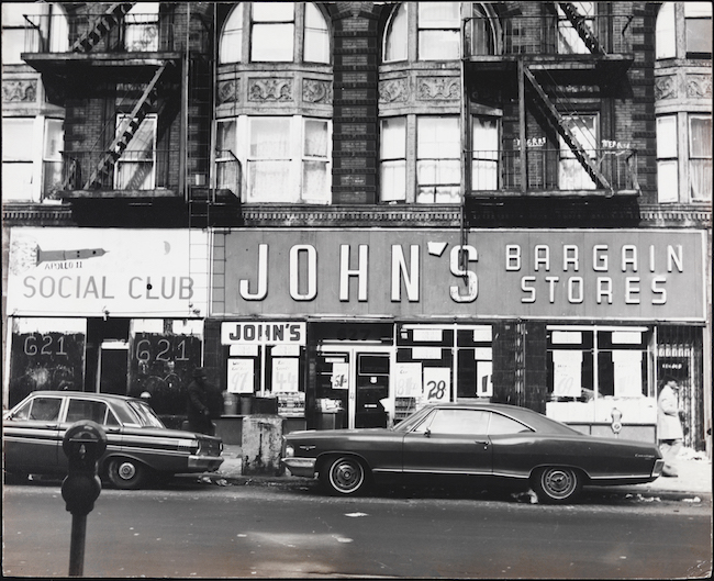 Vue de la rue Myrtle Avenue, Bed-Stuy, gros plan des devantures de magasins "John's Bargain Stores" et "Social Club" avec des voitures de ca. 1970 garé devant.