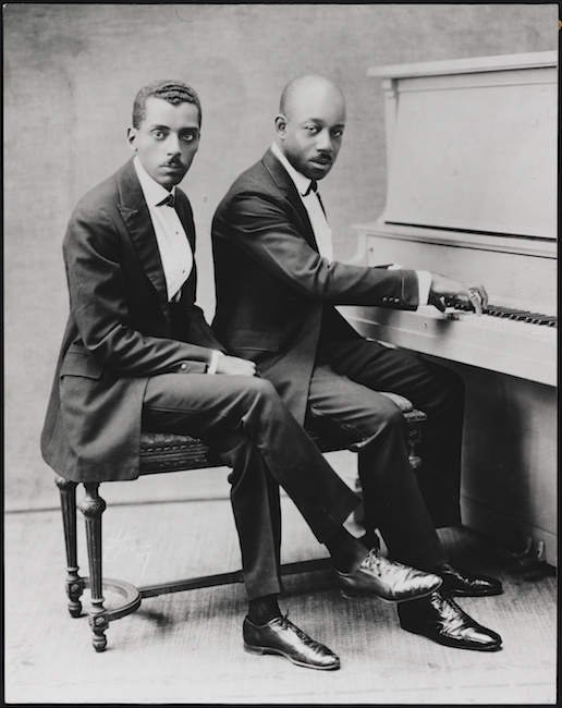정장을 입은 두 명의 아프리카 계 미국인 신사가 피아노 벤치에 앉아 오른쪽에 피아노가 있습니다. 오른쪽에있는 사람이 손을 키보드에 갖습니다.