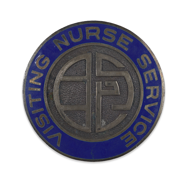 Épinglette en métal. Un dessin abstrait en métal uni au centre est entouré d'un anneau en émail bleu avec les mots "Visiting Nurse Services"