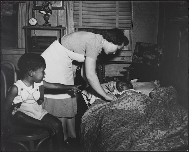 看護師が小さな子供をベッドに押し込み、椅子に座っている別の子供が見ています。