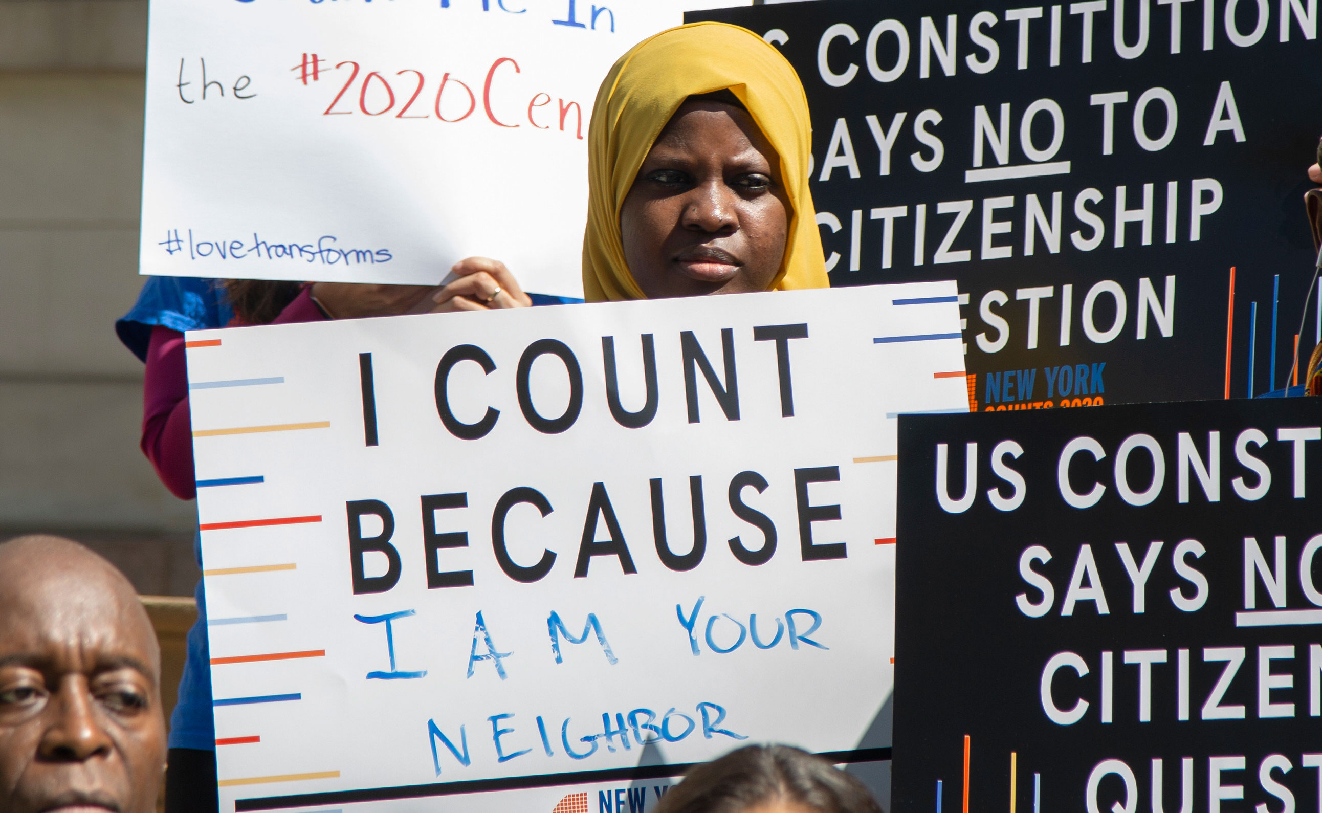 画像は、ニューヨーク市議会の記者会見で「私はあなたの隣人だからカウントする」というサインを持っている若者を示しています。