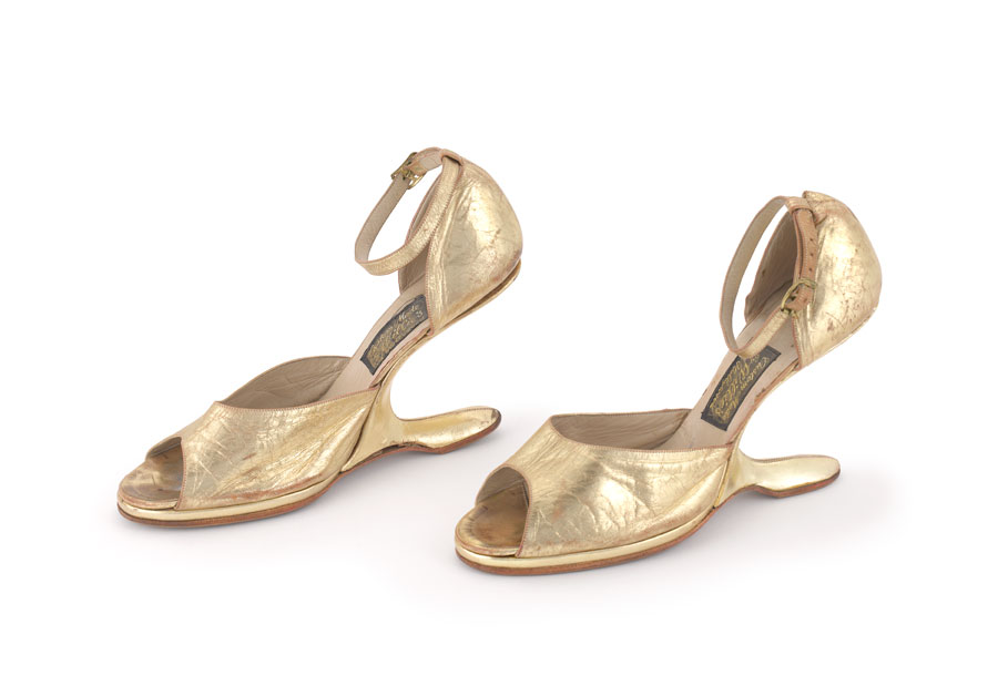 A pair of gold shoes, that are shaped like high heels. Rather than a heel, they have a piece that extends back from the base