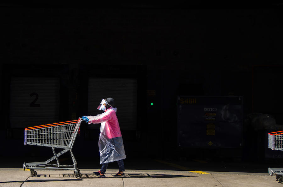 Una mujer con un protector facial y un abrigo de plástico empuja un carrito de compras.