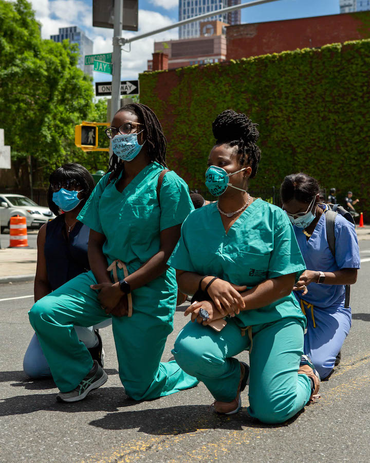 Quatro enfermeiras com uniforme e máscaras estão ajoelhadas em um estacionamento.