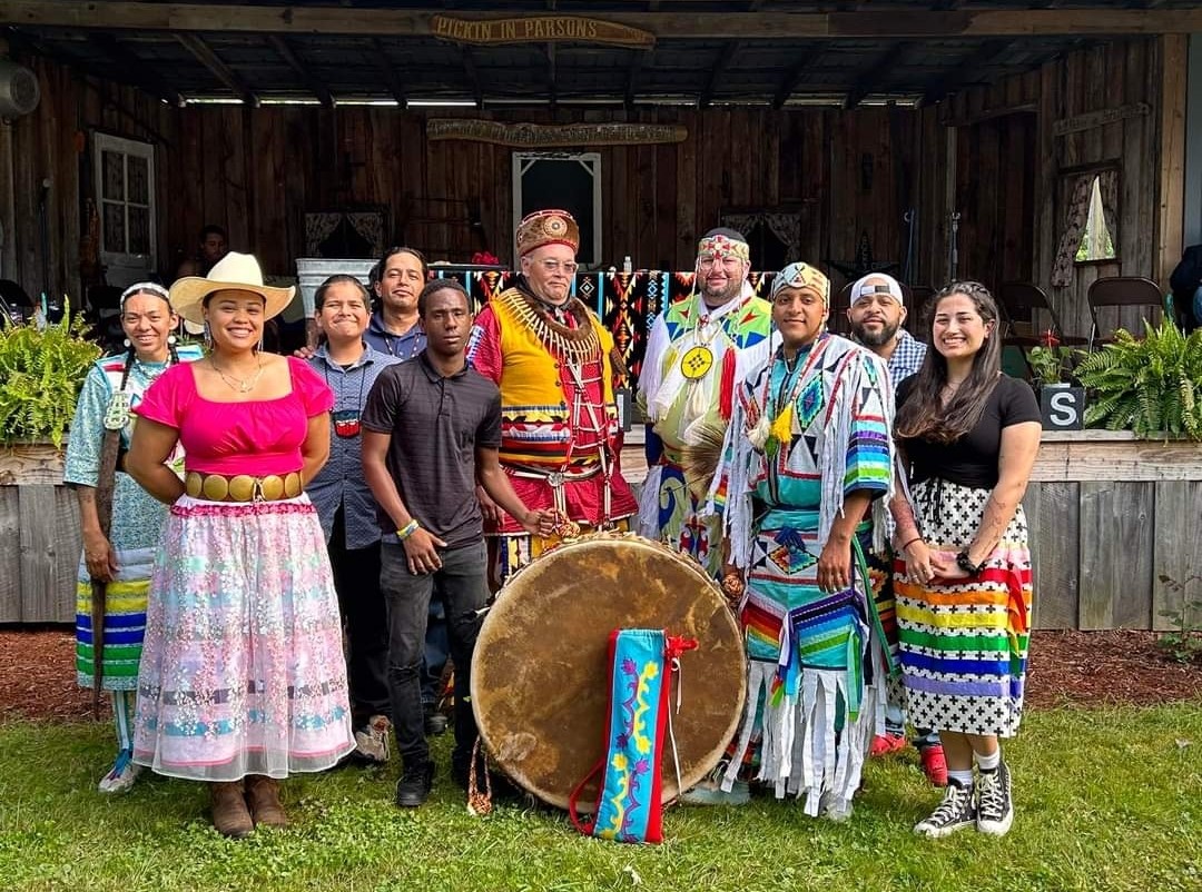Um grupo de homens e mulheres sorri para a câmera, vestindo uma mistura de roupas tradicionais dos nativos americanos e contemporâneas, ao redor de um tambor exibido em destaque.