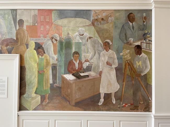 Photographie d'une fresque murale. Au centre se trouve une femme assise à un bureau avec une machine à écrire, une infirmière se tient devant.