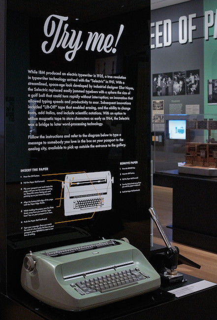 Vista de instalación de "Analog City: New York BC (Antes de las computadoras)", que muestra una pantalla con una máquina de escribir eléctrica que funciona y un texto que explica cómo usarla.