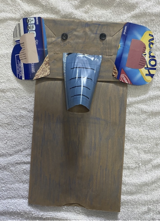 A lancheira de papel marrom se transformou em uma marionete projetada para parecer um elefante. Tem dois botões pretos para os olhos, duas orelhas feitas de um recipiente de comida de papelão azul e um tronco feito de um rolo de papel higiênico coberto de papel de revista azul.