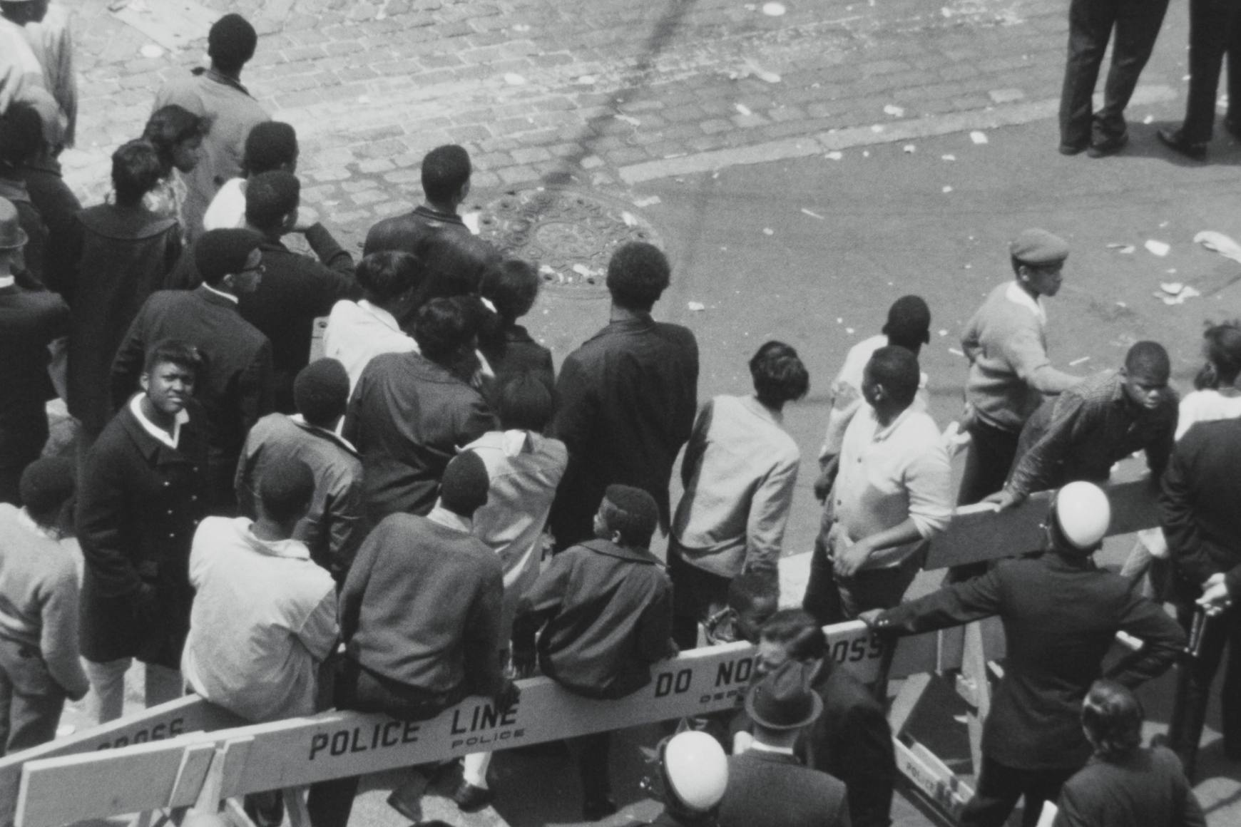 Uma visão panorâmica em preto e branco de um grupo de pessoas na rua. Barricadas policiais delineiam o espaço.