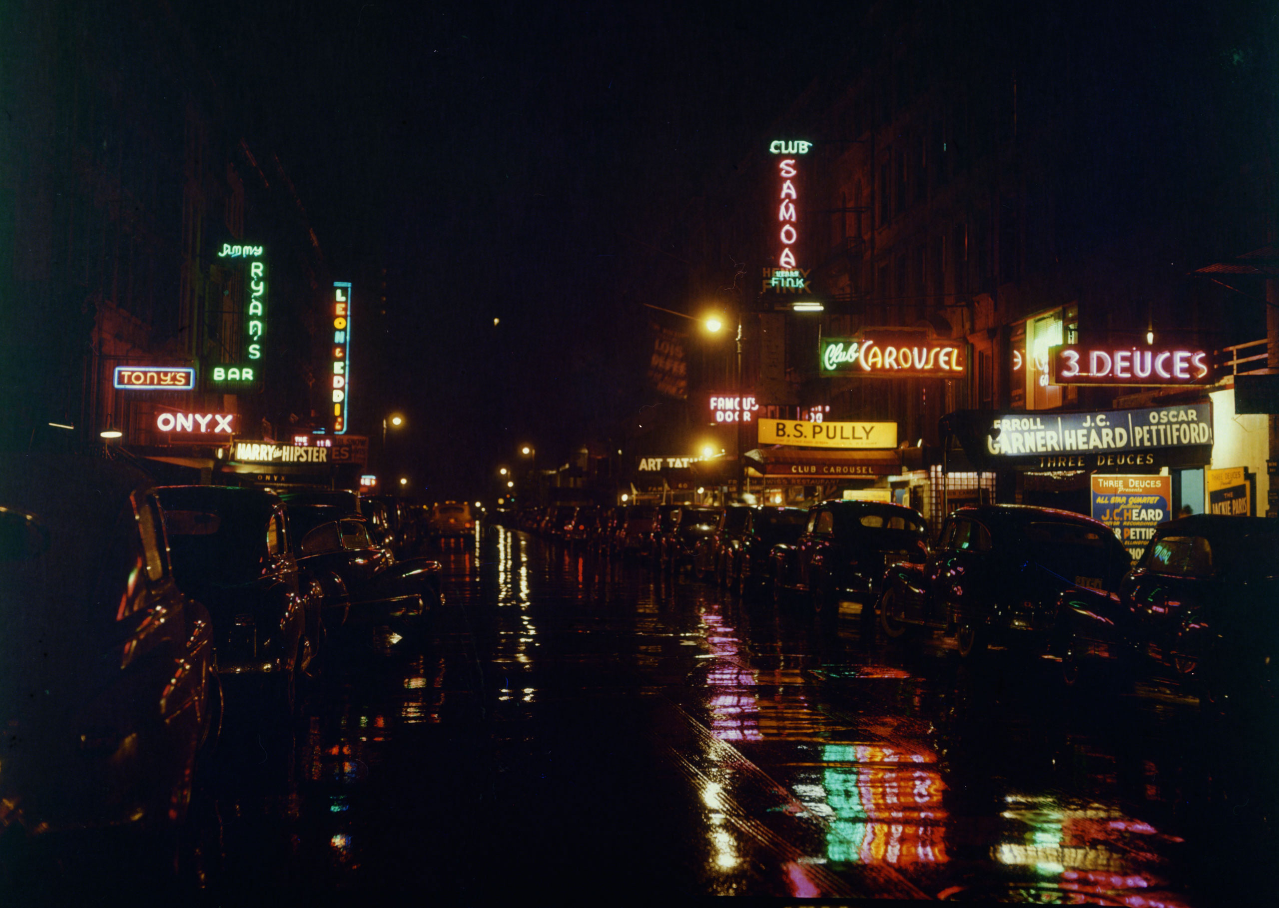 Estrada à noite, com sinais de néon refletindo na calçada molhada e carros estacionados em ambos os lados da rua