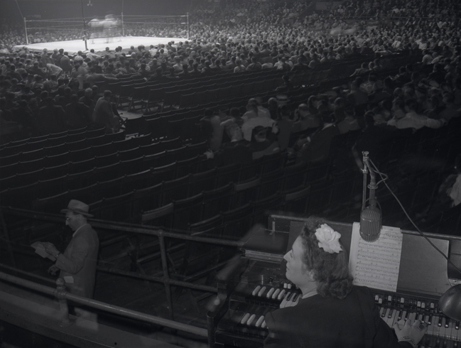 L'organiste des sports Gladys Gooding est assis à un orgue au Madison Square Garden avec un ring de boxe et un public à l'arrière-plan.