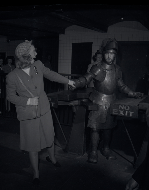 Un hombre con una armadura de metal camina por un torniquete del metro mientras una mujer con un traje le toma la mano y lo conduce.