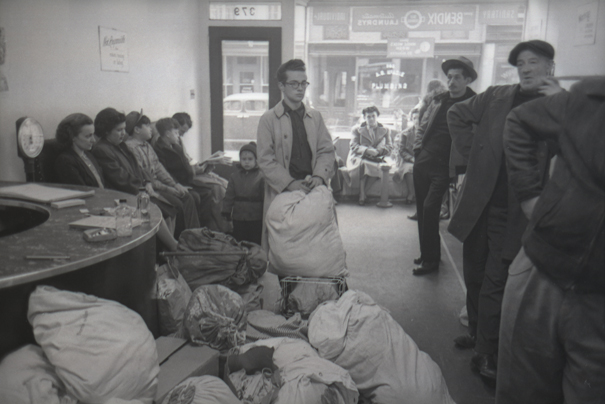 Varios hombres y mujeres se paran y se sientan en una lavandería de Greenwich Village con bolsas de lavandería y lavadoras sentadas frente a ellos.