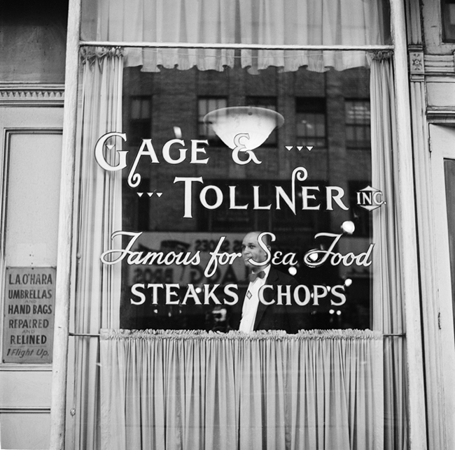 Exterior of Gage and Tollner restaurant. The lettering on the window reads “Gage & Tollner Inc. Famous for Sea Food, Steaks, Chops.” A waiter is visible through the window.