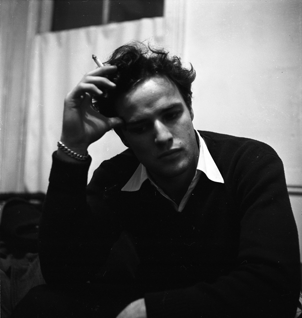 演员马龙·白兰度（Marlon Brando）坐在室内，穿着一件毛衣，拿着烟。