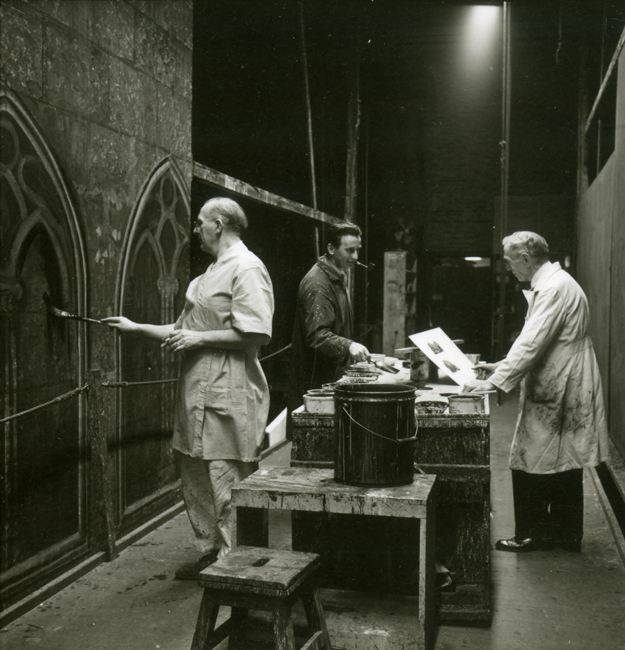 Trois hommes en blouses et tabliers tiennent des pinceaux et des papiers pendant qu'ils peignent des ensembles pour le Metropolitan Opera avec des tables de travail et des seaux de peinture à côté d'eux.