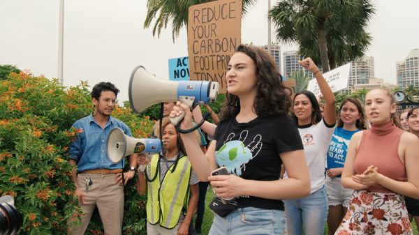 Jamie Margolin détient un mégaphone bleu et blanc dans lequel elle parle activement. Une foule de jeunes suit derrière elle en criant et en tenant des pancartes qui disent "Réduisez votre empreinte carbone aujourd'hui."