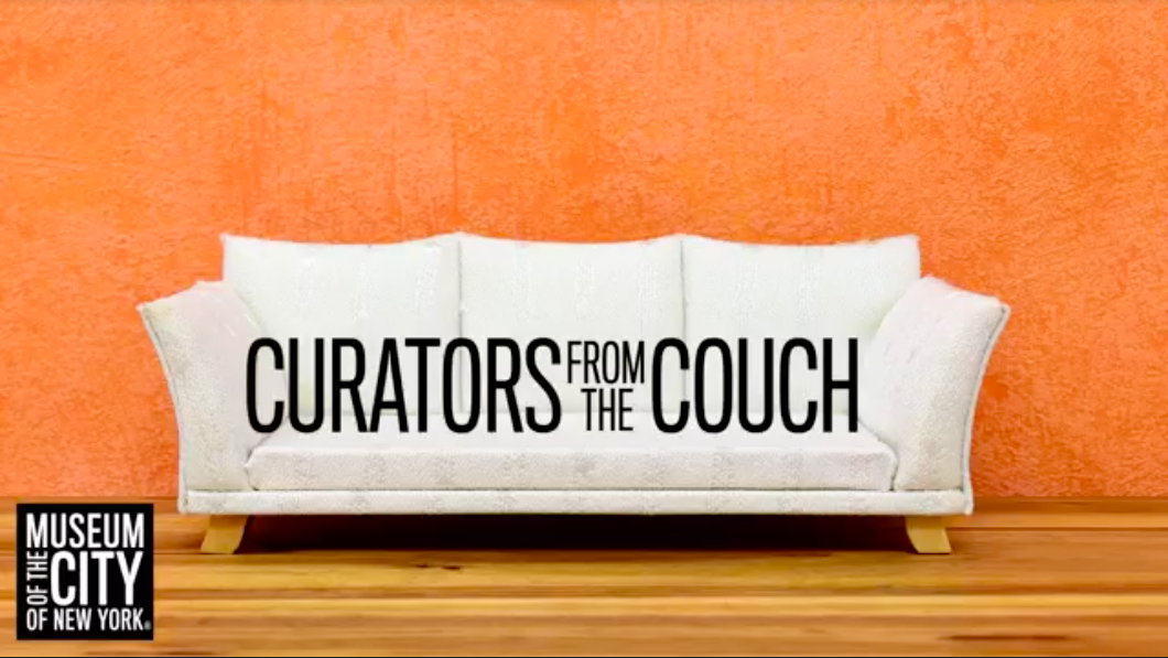 Sofá blanco contra una pared pintada de naranja y piso de madera. El texto en el sofá dice "Curadores desde el sofá" en letras negras.