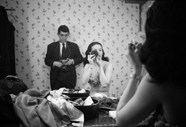 一个女人坐在服装的镜子，化妆表演。 一个男人用相机靠在墙上。