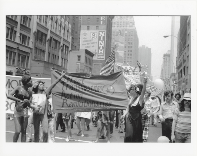 Photographie en noir et blanc représentant un défilé lors de la Journée de libération de Christopher Street. Trois personnages, devant, tiennent une bannière pour STAR, ou Street Transvestite Action Revolutionaries.