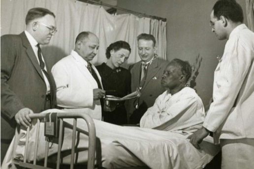 Fotografia em preto e branco do dr. Lyndon M. Hill, do dr. Louis T. Wright, do dr. Myra Logan, do dr. Aaron Prigot e de um funcionário não identificado do hospital em pé ao lado da cama de uma paciente afro-americana não identificada.