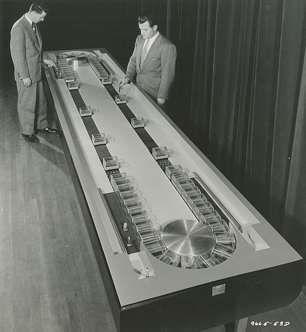 Two men in suits examining a working model of conveyor subway system.