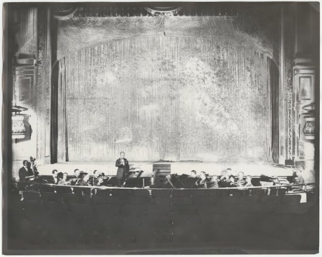 剧院舞台的图像，画着窗帘，乐团坐在前面，指挥家站着。
