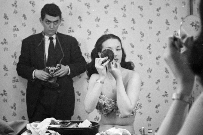 Tomada no reflexo de um espelho, uma mulher usa um espelho compacto para aplicar maquiagem, enquanto o fotógrafo fica atrás dela assistindo