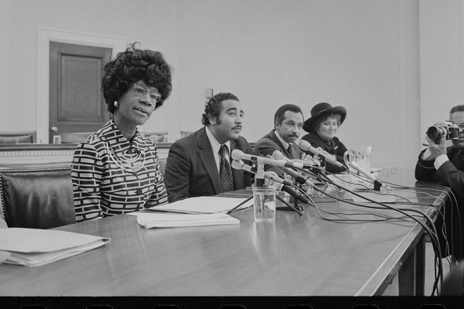 照片显示众议员雪莉·奇泽姆 (Shirley Chisholm)、众议员帕伦·米切尔 (Parren Mitchell)、众议员查尔斯·兰格尔 (Charles Rangel) 和众议员贝拉·阿布祖格 (Bella Abzug) 坐在带麦克风的桌子旁