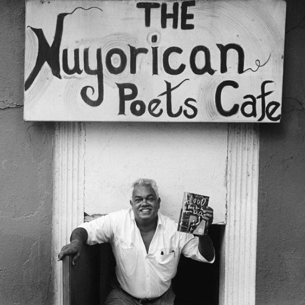 Un hombre, el poeta Miguel Algarín, sonríe y sostiene un libro bajo un cartel que dice: "El Café de los Poetas Nuyorican".