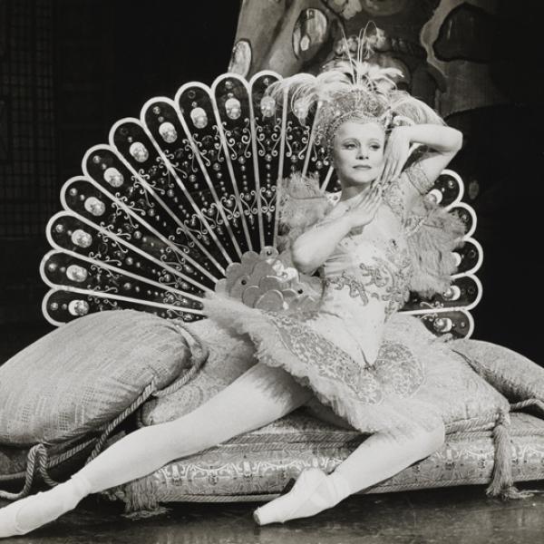 Una bailarina en traje se reclina sobre cojines con una estilizada cola de pavo real desplegada detrás de ella.